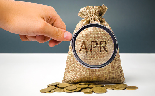 A hand points to a money bag with the word Apr. This is a financial fee, expressed as an annual rate. Applicable to loan, mortgage, credit card. Commercial money investment. APR. Percentage Rate