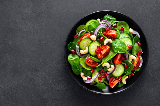 istock Spinach salad with fresh cucumbers, tomato, onion, pomegranate, sesame seeds and cashew nuts on black background. Healthy vegan food. Top view 1129700921