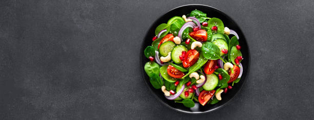 istock Spinach salad with fresh cucumbers, tomato, onion, pomegranate, sesame seeds and cashew nuts on black background. Healthy vegan food. Top view. Banner 1129700961