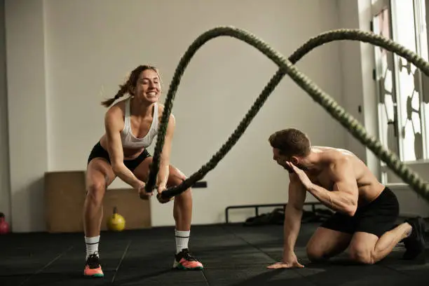 Dedicated sportswoman exercising with battle rope while coach is encouraging her during cross training in health club.