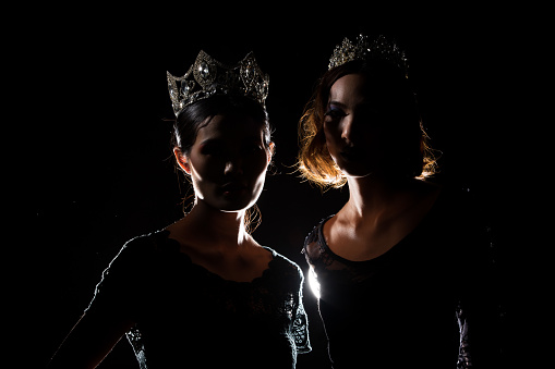 Two Silhouette Shadow Back Rim Light of Miss Pageant Beauty Queen Contest with Silver Diamond Crown stand together, studio lighting dark black background, multinational asian and caucasian models
