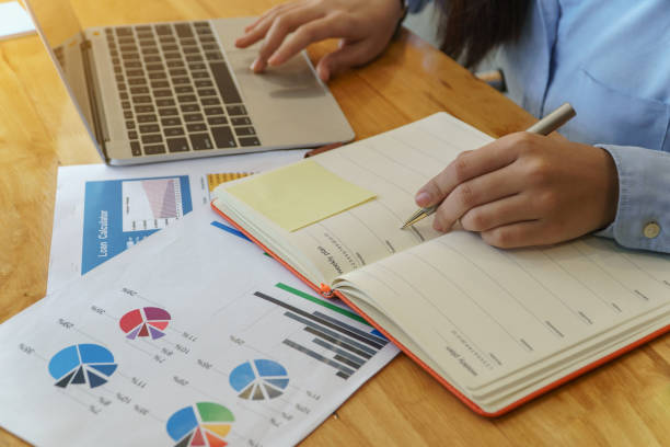 istock Young business woman sitting at table and taking notes in notebook. Loan investment documents. 1129779823