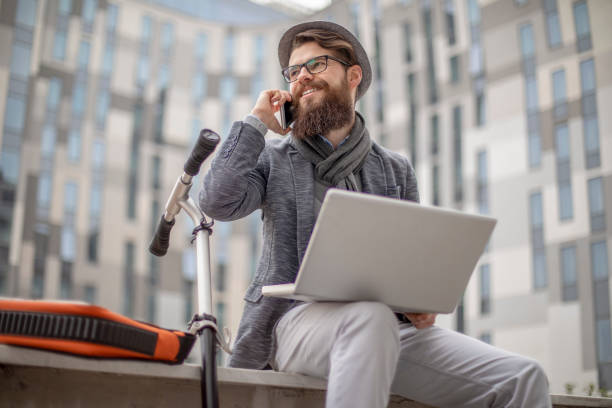 istock Young bearded businessman with kick scooter, lap top and hat in outer environment 1129781676
