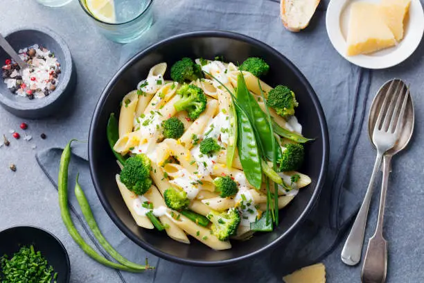 Pasta with green vegetables and creamy sauce in black bowl on grey stone background. Top view