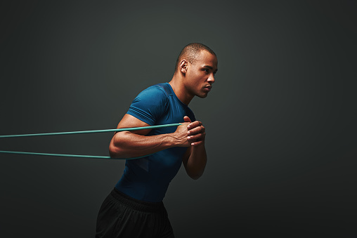 Isolated studio portrait of clean shaven young dark skinned male athlete with strong muscular shoulders working out using resistance elastic band, strengthening arm muscles over dark background. Side view