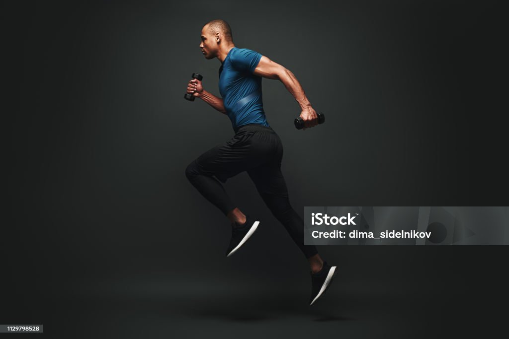 Everything you ve ever wanted is on the other side of fear. Dark skinned sportsman jumping over dark background. He is ready to run Full length portrait of a healthy muscular sportsman jumping isolated over dark background, with dumbbells in his hands. Dynamic movement. Side view Exercising Stock Photo
