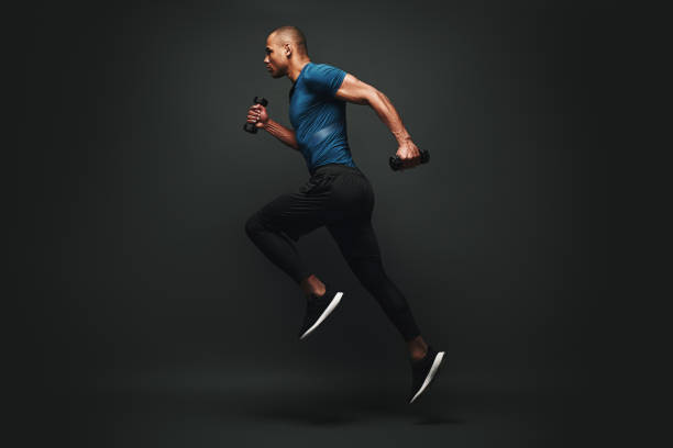 Everything you ve ever wanted is on the other side of fear. Dark skinned sportsman jumping over dark background. He is ready to run Full length portrait of a healthy muscular sportsman jumping isolated over dark background, with dumbbells in his hands. Dynamic movement. Side view gym photos stock pictures, royalty-free photos & images