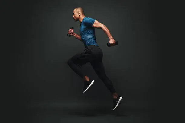 Full length portrait of a healthy muscular sportsman jumping isolated over dark background, with dumbbells in his hands. Dynamic movement. Side view