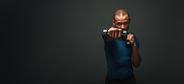 Sport, bodybuilding, fitness and people concept - man with dumbbells flexing muscles and looking at the camera isolated on dark background. Long shot