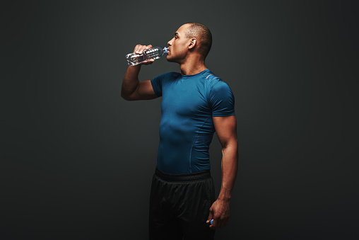 Exhausted dark skinned athlete in sportswear drinking water from plastic bottle after training. Studio shot isolated over dark background