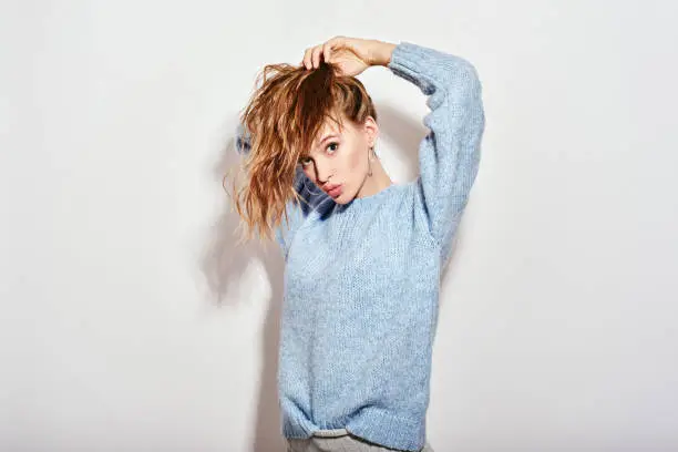 Portrait of young beautiful girl with brown hair, full lips, wearing blue sweater. She is fixing her ponytail, looking flirty at camera, isolated over white background