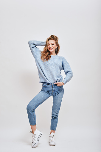 Full-length portrait of playful, vivacious girl wearing blue sweater, jeans, converse, fixing her hair, while smiling happily at camera, isolated over white background.