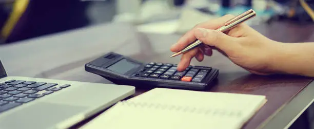 Photo of close up accountant employee man hand using pen for writing on paper check and press on calculator for calculate coast estimate monthly of company's profit and typing keyboard on laptop for information at office , business concept