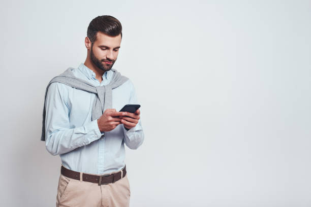 istock Very busy. Handsome young man in casual wear using his smart phone while standing on a grey background. Digital concept 1129816867