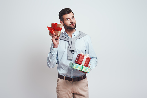 Buy gifts in advance. Good looking and smilling man is holding gifts on a grey background. Close-up portrait