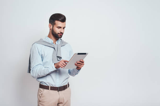 istock Very busy. Stylish bearded man in a casual wear is using his digital tablet for work while standing on a grey background. Digital concept 1129816973