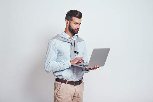 Modern technologies. Successful young man with a beard using his laptop while standing against grey background. Digital concept