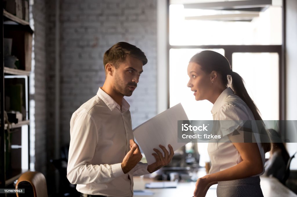 Diverse employees argue over financial report in office Millennial diverse employees standing holding financial paperwork, disputing over statistics or contract terms, man and woman have misunderstanding in office, argue or quarrel on document or report Arguing Stock Photo