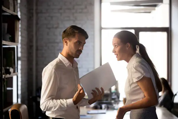 Millennial diverse employees standing holding financial paperwork, disputing over statistics or contract terms, man and woman have misunderstanding in office, argue or quarrel on document or report