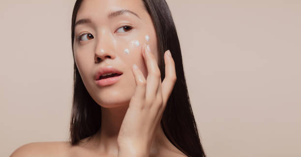Applying moisturizer to her face Close up of a youthful female model applying moisturizer to her face. Young korean woman applying moisturizer cream on her pretty face against beige background. asian american model stock pictures, royalty-free photos & images