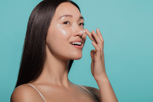 Close up of female model applying moisturizer cream to her nose. Woman applying moisturizer cream on her face against blue background.