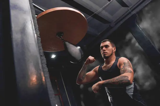 Photo of Born to win. Muscular tattooed athlete in sports clothing training hard on punching speed bag to become a champion, boxing gym interior