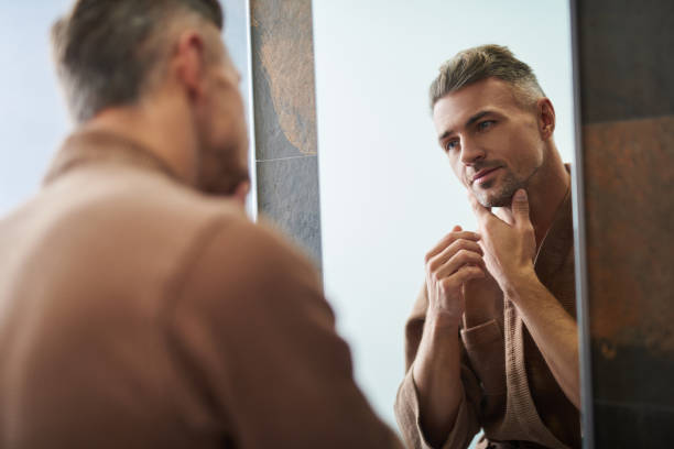 istock Handsome unshaven man looking in the mirror at home bathroom 1129826881