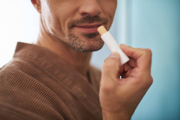 Young man using hygienic lipstick on blue background