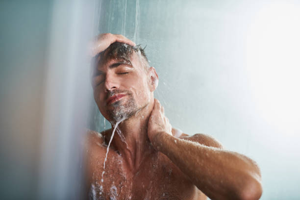 beau jeune homme avec des yeux fermés debout sous les gouttes d’eau et souriant - prendre un bain photos et images de collection