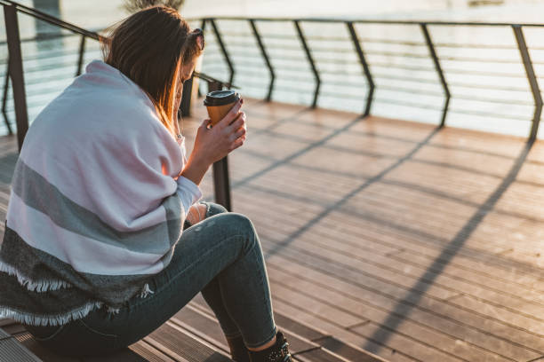 Solitude or depression concept. Millennial dealing with problems and emotions. Sad woman standing by the water during sunset. Sad and depressed woman sitting alone