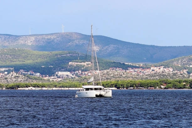 un moderno crucero de vela catamarán con un aparejo de bermudas sloop-tipo va más allá de la verde costa de la riviera croata en un día soleado de verano. mar adriático de la región mediterránea. distrito de dalmacia - riff fotografías e imágenes de stock