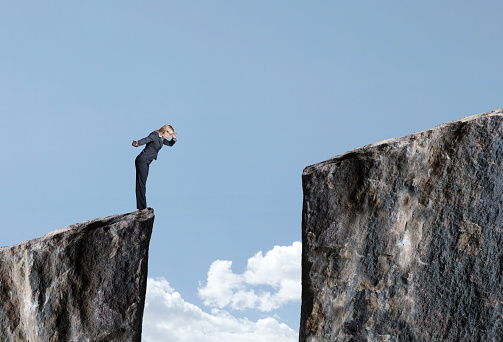 A businesswoman stands at the edge of a cliff and looks down at the large gap that prevents her from proceeding forward.