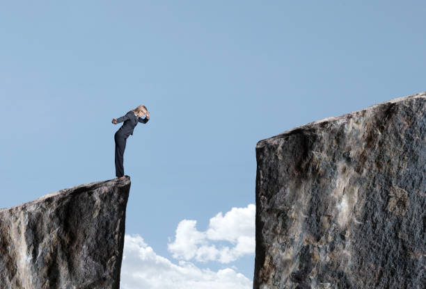 Businesswoman Looks Down At Large Gap Between Two Cliffs A businesswoman stands at the edge of a cliff and looks down at the large gap that prevents her from proceeding forward. gender stereotypes stock pictures, royalty-free photos & images
