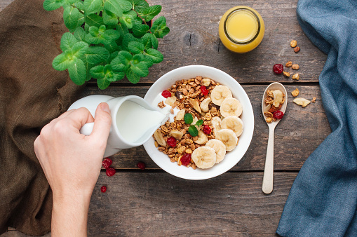 Close up of woman pouring milk into bowl with granola on rustic wooden background. Top view, flat lay.