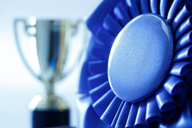 A close up of the rosette of a blue ribbon in front of a trophy out of focus in the background.