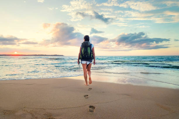 istock Woman with backpack walk on the ocean sand beach at sunset time 1129909413