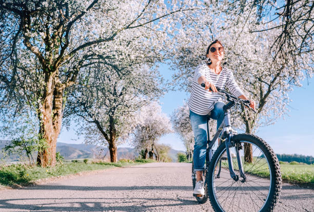 feliz sonriente mujer monta una bicicleta en la carretera bajo los árboles de flor. primavera viene imagen de concepto. - comming fotografías e imágenes de stock