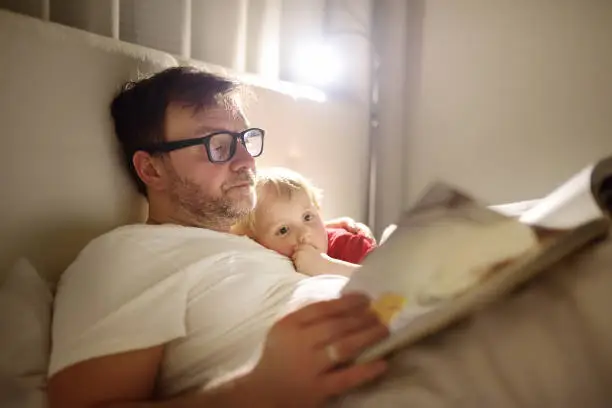 Photo of Father reading bedtime stories to child. Dad putting son to sleep