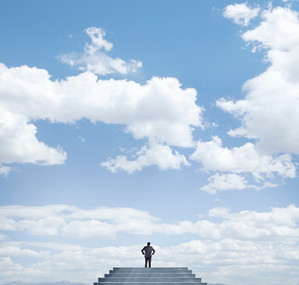 A businessman has climbed a set of stairs and is dwarfed by the cloud filled sky in front of him.
