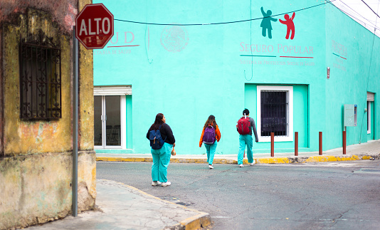 Merida, Yucatan, Mexico: Three young people wearing turquoise medical scrubs and backpacks walk across a street in downtown Merida, toward a bright turquoise building, housing government medical services.