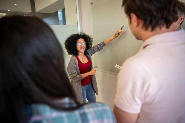 istock Beautiful black female student explaining something to classmates pointing at glass board 1129999753