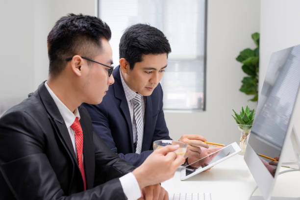 retrato cintura arriba de dos empresarios guapos de traje sentado en la mesa con el portátil en el interior de la oficina, hablando y mirando el uno al otro mientras se discuten un nuevo proyecto - waistup fotografías e imágenes de stock