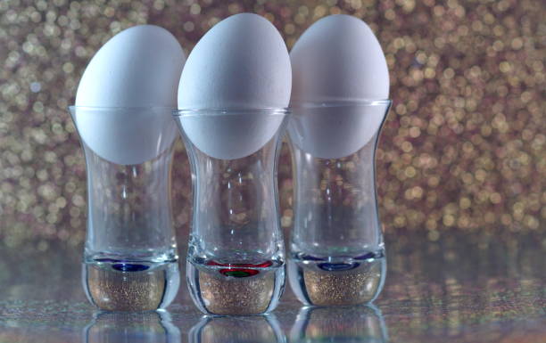 Three white eggs in small glasses with mirror reflection on a gold glittering background abstract conceptual art photography