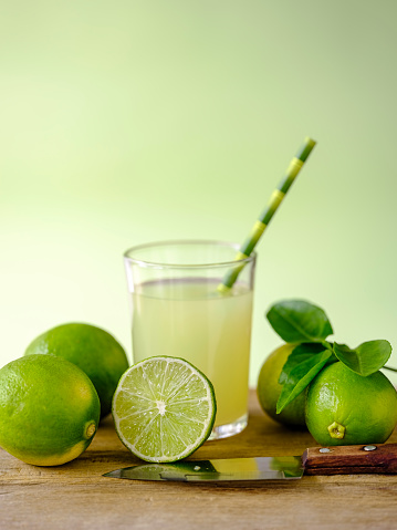 Group of limes next to a drinking glass with fresh lime juice and a paper drinking straw on a cutting board with a small kitchen knife, some limes are on the table, some cut in cross section, set on an old weathered wood table background, shallow depth of field with focus on the lime in cross section, good copy space above the image.