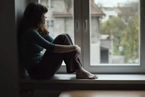 Photo of Sad young woman sitting on the window, watching out