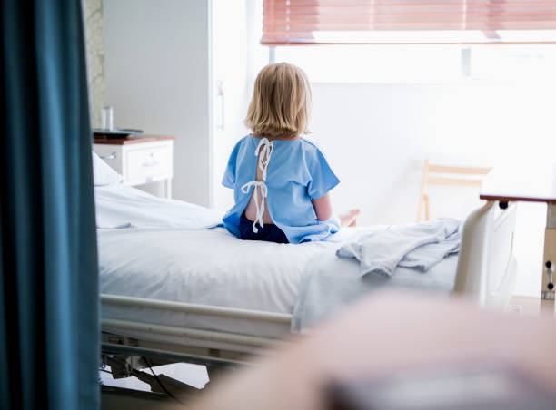 widok z tyłu chorej dziewczyny siedzącej na łóżku w szpitalu - examination gown zdjęcia i obrazy z banku zdjęć