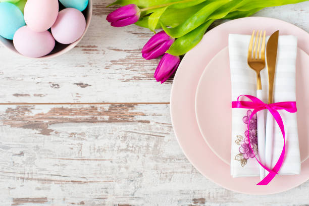 istock Easter table setting, flatlay with pastel colored Easter eggs, event decoration. Pink dishes and a gold fork and a spoon on a light rustic wooden background. Beautiful purple spring tulip flowers 1130086236