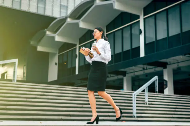 Busy Asian businesswoman in eyeglasses walking to work at rush hour in front office holding smartphone- coffee cup and business file in morning-sun flare- woman office worker and businesswoman concept