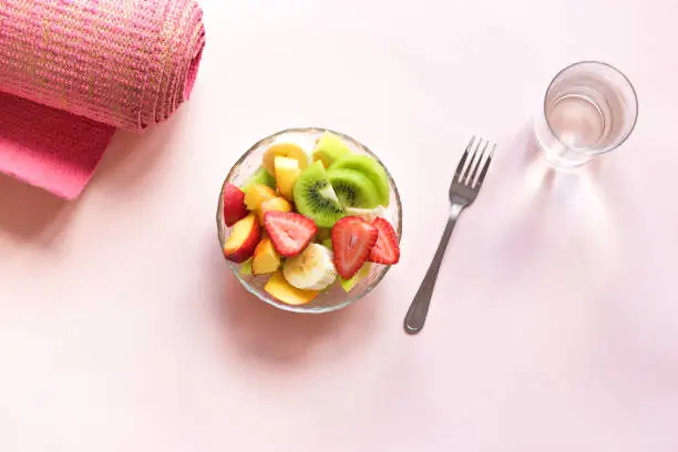 Feminine Sport and Healthy Lifestyle Concept. Pink yoga mat and Fruit and Berries Salad in bowl for snack or breakfast on pink pastel background, copy space.