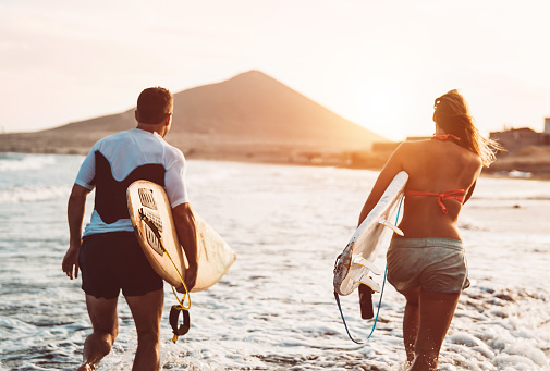 Happy surfer couple running with surfboards along the sea shore - Sporty people having fun going to surf together at sunset - Extreme surfing sport and youth relationship lifestyle concept